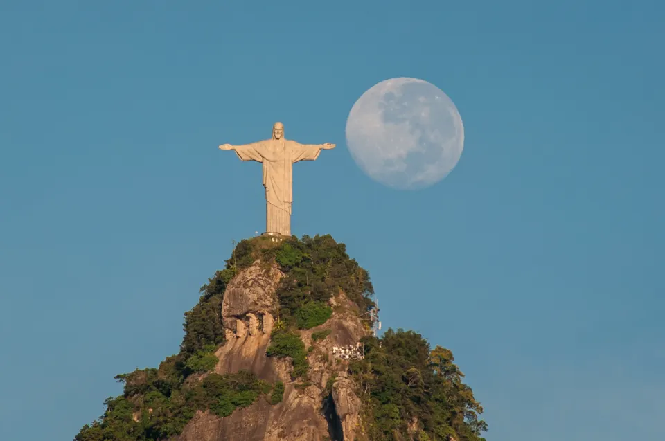 Christ the Redeemer on the Corcovado summit beside a full moon in a blue sky