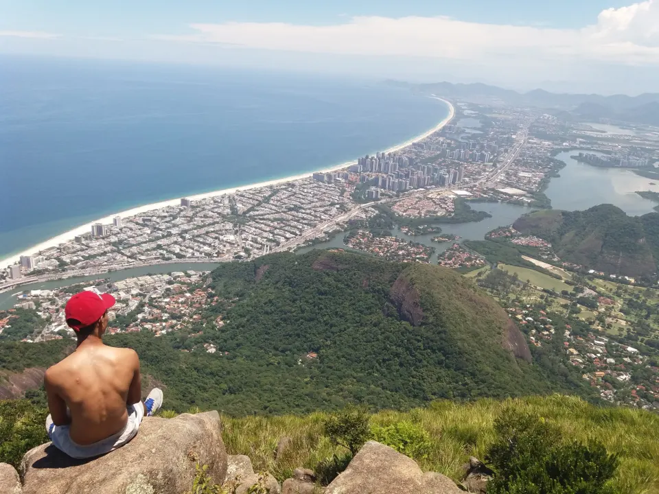 Hiker resting on the Pedra da Gávea summit overlooking Barra da Tijuca beach
